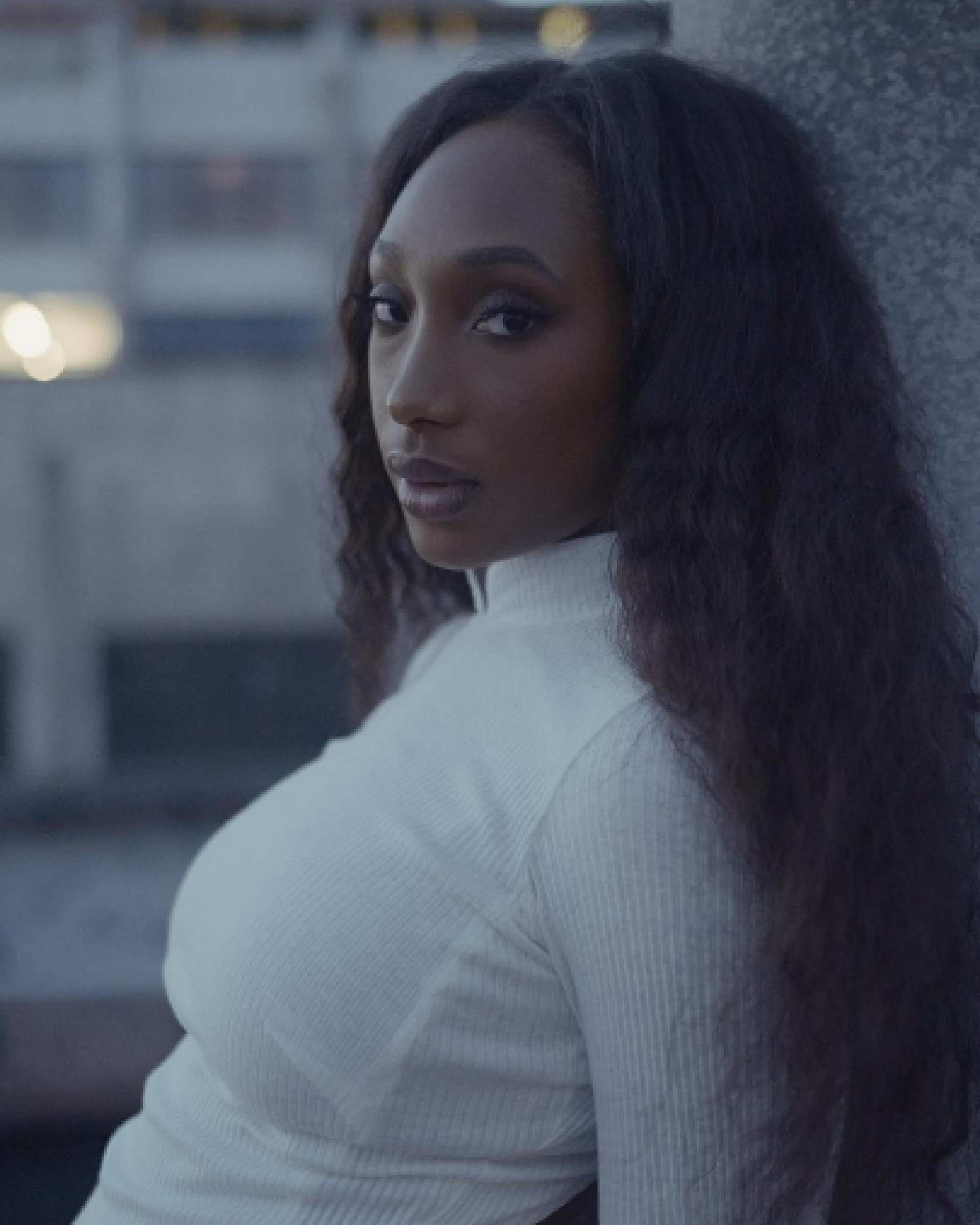 Woman in a white high-neck top leaning against a wall with blurred background